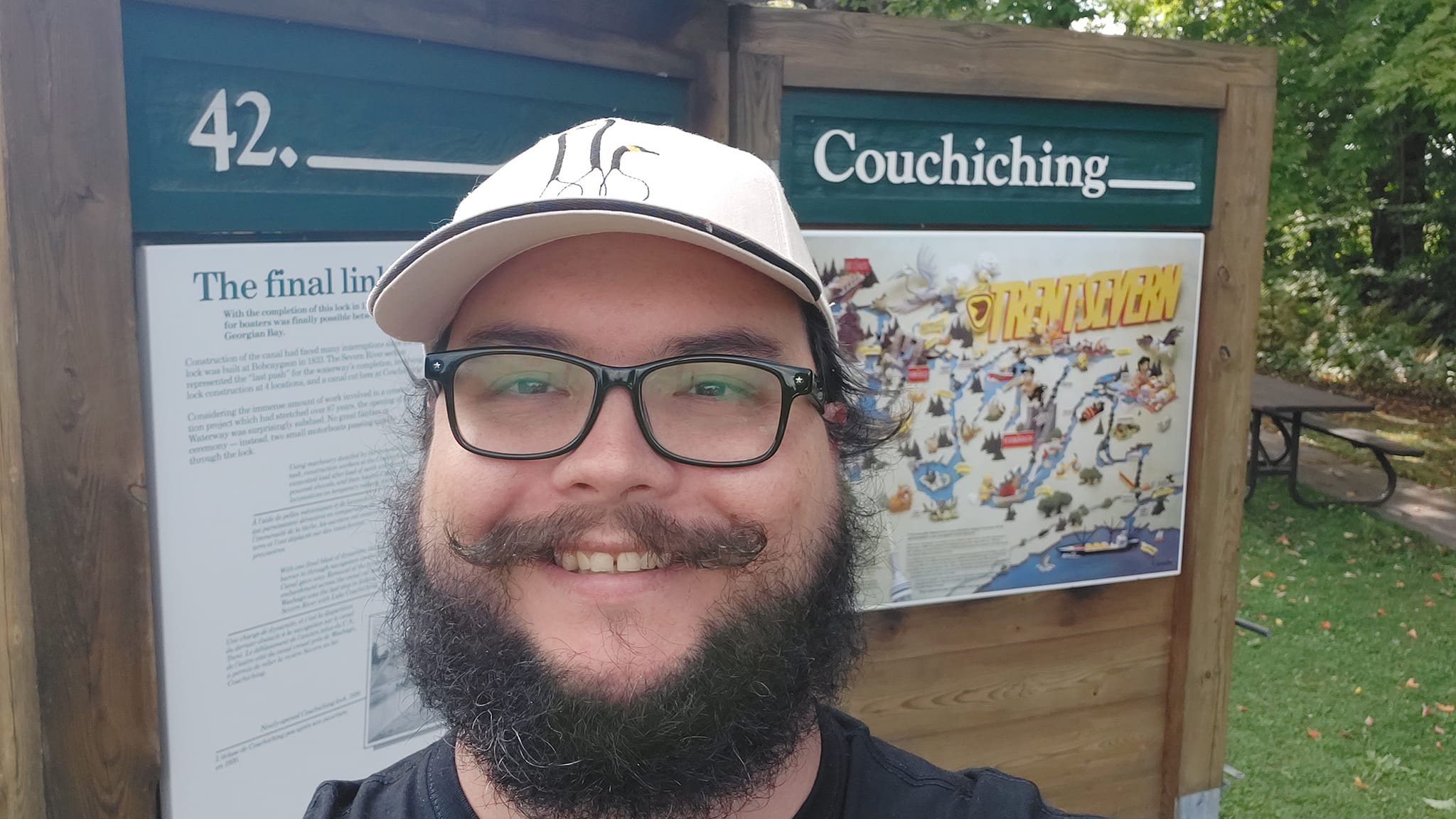 A man with short hair and bushy facial hair stands in front of a display board that reads "42. Couchiching", it is a sign for a lock station on a waterway